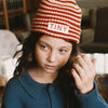 A young girl wearing a red and white striped knit beanie with 'TINY' embroidered on the front, paired with a blue cardigan featuring a pink mushroom design, in a softly lit indoor setting.