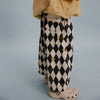 A close-up of a child's lower body wearing black and cream harlequin patterned pants with yellow buttons, a beige top, and beige clogs, set against a solid light blue-gray background.